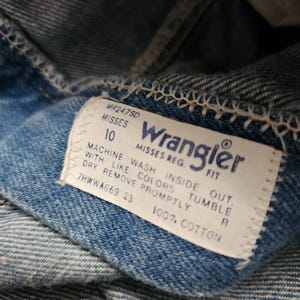 May include: A blue denim garment featuring a white rectangular interior tag with the Wrangler logo in blue text. The tag includes the text MR247SD, MISSES 10, MISSES REG. FIT, 100% COTTON, and care instructions: MACHINE WASH INSIDE OUT, WITH LIKE COLORS, TUMBLE DRY, REMOVE PROMPTLY.