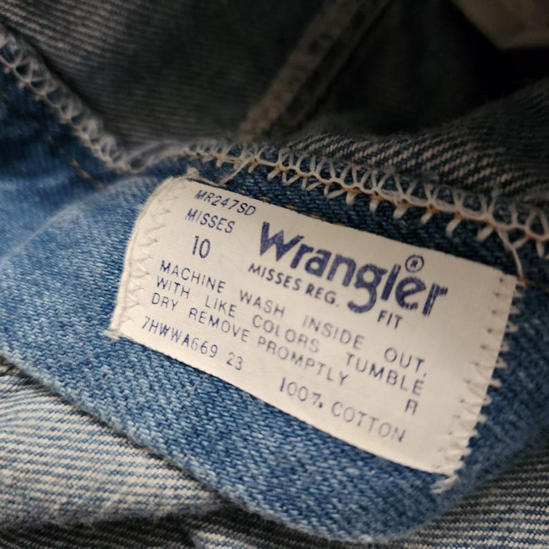 May include: A blue denim garment featuring a white rectangular interior tag with the Wrangler logo in blue text. The tag includes the text MR247SD, MISSES 10, MISSES REG. FIT, 100% COTTON, and care instructions: MACHINE WASH INSIDE OUT, WITH LIKE COLORS, TUMBLE DRY, REMOVE PROMPTLY.