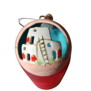 Könnte beinhalten: Keramik-Ornament mit einem weißen Gebäude im Pueblo-Stil mit roten Akzenten, einer Leiter und blauen Fenstern. Das Ornament ist in einem rot-beigen tropfenförmigen Rahmen mit einer Metallschlaufe zum Aufhängen eingefasst.