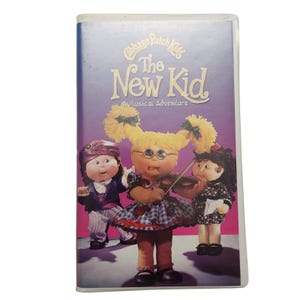 Könnte beinhalten: Ein VHS-Band mit den Cabbage Patch Kids in einem musikalischen Abenteuer namens "The New Kid". Das Cover zeigt drei Cabbage Patch Kids-Puppen, eine spielt Geige, eine hält ein Musikbuch und eine trägt einen lila Hut.