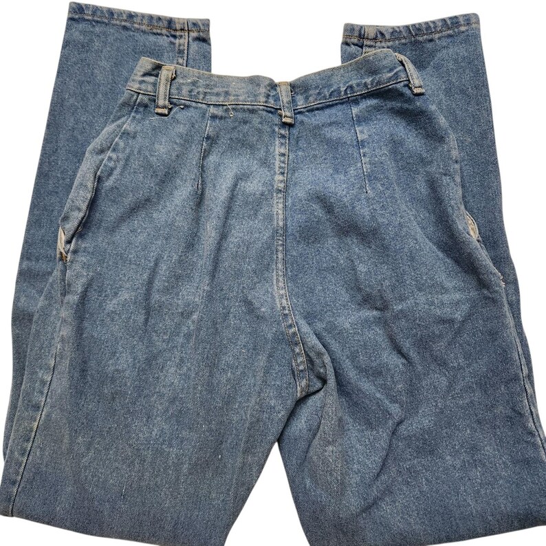 May include: High-waisted blue denim jeans featuring a classic five-pocket design, a button and zip fly closure, belt loops, and vertical pleats at the front waistline. The fabric has a medium wash finish with visible stitching throughout.