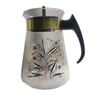 Puede incluir: Una cafetera de vidrio transparente con asa negra y ribete dorado. La cafetera tiene un diseño floral con hojas y flores negras y doradas.