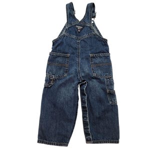 May include: Dark blue denim overalls with multiple pockets, including two front pockets, two back pockets, and a small bib pocket. The overalls have adjustable straps and button closures. The brand name "OshKosh B'gosh" is visible on the bib.