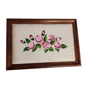 Könnte beinhalten: Eine gerahmte Stickerei mit rosa Rosen und grünen Blättern. Das florale Design ist auf einem cremefarbenen Stoff zentriert, der in einem rechteckigen Holzrahmen eingefasst ist. Die Stickerei zeigt detaillierte Stiche.