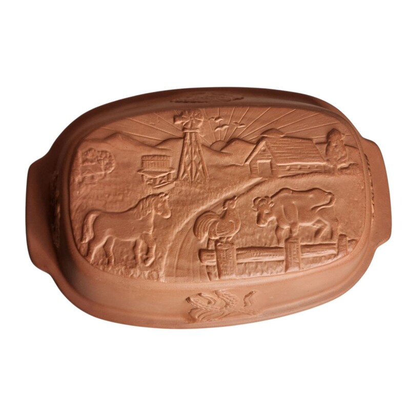 May include: A terracotta-colored, oval-shaped baking dish with a detailed farm scene carved into the surface. The scene includes a horse, cow, rooster, windmill, barn, and wheat stalks. The dish has two handles.