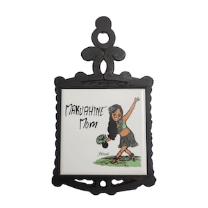Può includere: Sottopentola in ghisa nera con un inserto in ceramica bianca. La piastrella presenta una ballerina di hula in stile cartone animato con il testo "Makuahiine Mom" e "Hawaiian".