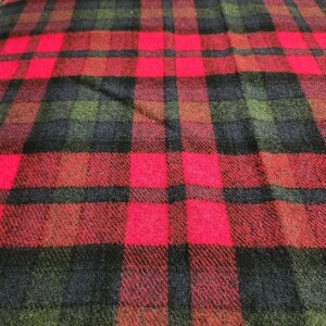 May include: A close-up view of a red, black, and green plaid fabric. The pattern consists of large squares in red, black, and green, forming a traditional design. The fabric looks to be woven, possibly wool or a similar textile.