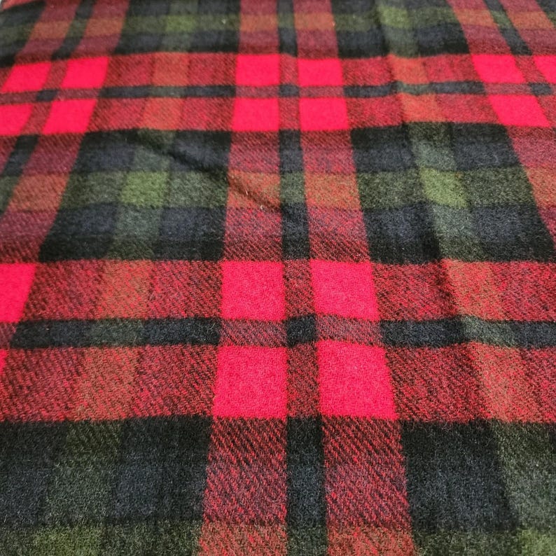 May include: A close-up view of a red, black, and green plaid fabric. The pattern consists of large squares in red, black, and green, forming a traditional design. The fabric looks to be woven, possibly wool or a similar textile.