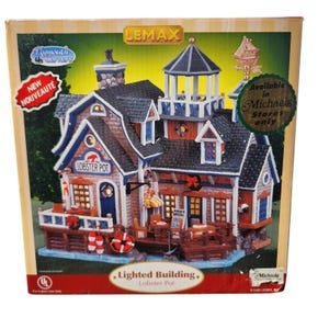 Może przedstawiać: Podświetlany miniaturowy budynek Lobster Pot z ciemnoszarym dachem, czerwonymi i niebieskimi akcentami oraz znakiem "Lobster Pot". Budynek posiada mały taras ze stolikami i krzesłami. Na pudełku napisano "Dostępne tylko w sklepach Michaels".