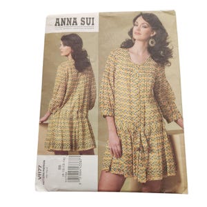 May include: Anna Sui V1177 sewing pattern. The pattern features a dress with a V-neck, long sleeves, and a tiered skirt. The dress is shown in a yellow and blue patterned fabric. The pattern includes sizes 8-14.