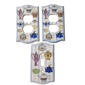 May include: Three rectangular ceramic wall plates featuring a light blue and white vertical striped pattern decorated with various colorful teapots, coffee pots, and cups. Two plates have double outlet openings, and one has a single toggle switch.