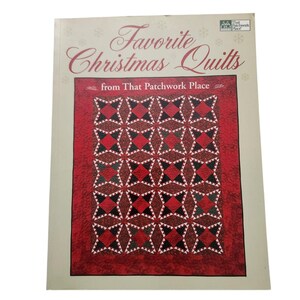 Puede incluir: Un libro titulado "Favorite Christmas Quilts" de That Patchwork Place. La portada presenta un diseño de edredón rojo y negro con patrones de estrellas y diamantes. El libro tiene un borde beige claro.