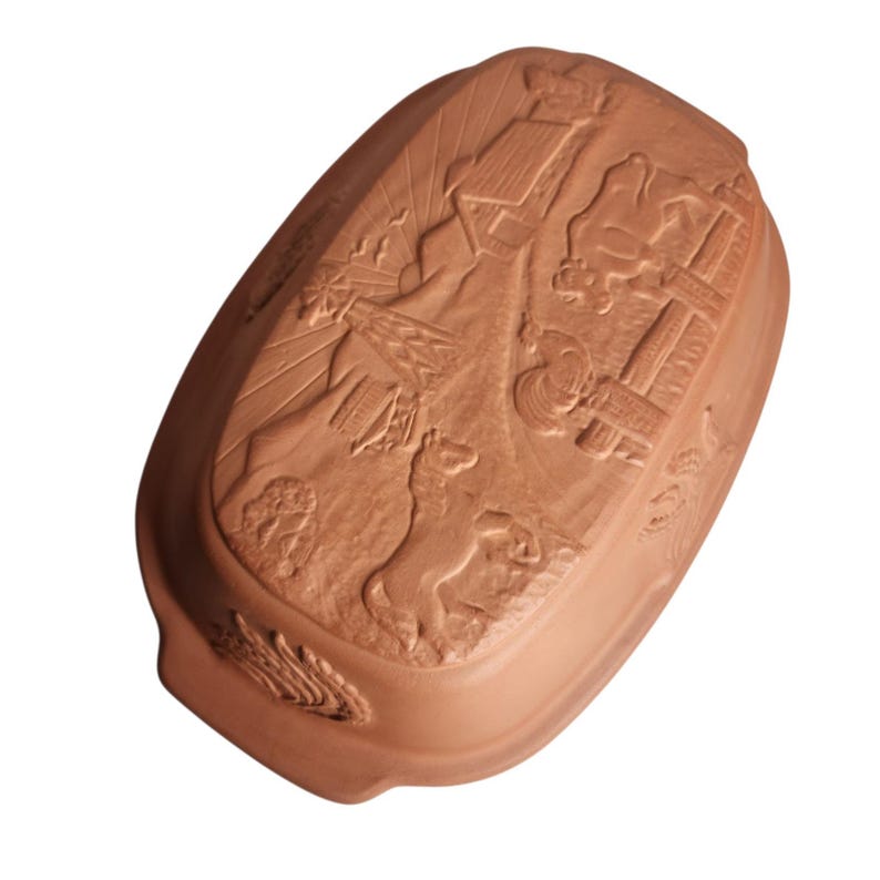 May include: A terracotta-colored, rectangular baking dish with a detailed relief design. The relief depicts a scene with figures, buildings, and a dog. The dish has a slightly curved shape and is suitable for baking.