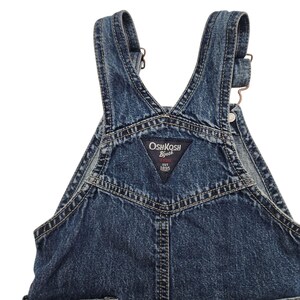 May include: Close-up of blue denim overalls. The overalls have adjustable straps, metal hardware, and a triangular patch with the "OshKosh B'gosh" logo. The denim has a faded, worn appearance.