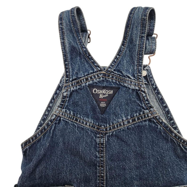 May include: Close-up of blue denim overalls. The overalls have adjustable straps, metal hardware, and a triangular patch with the "OshKosh B'gosh" logo. The denim has a faded, worn appearance.