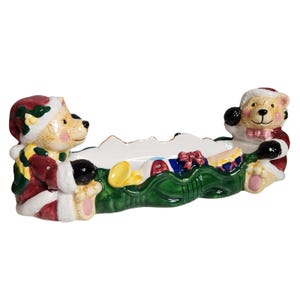 May include: A ceramic Christmas serving dish featuring two teddy bears in Santa outfits. The bears flank a green garland filled with wrapped gifts and a golden horn. The dish is white with a gold trim and is perfect for holiday treats.