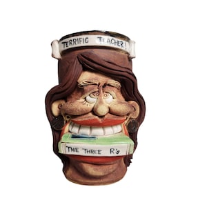 May include: A brown ceramic mug shaped like a woman's face with brown hair. The mug has a wide smile with white teeth and is holding a book that says "The Three R's". The mug has a banner that says "Terrific Teacher!"
