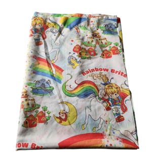 May include: A white fabric sheet featuring a repeating pattern of Rainbow Brite characters riding white horses, colorful rainbows, small houses, and yellow crescent moons. The text Rainbow Brite is printed in red letters across the fabric surface.
