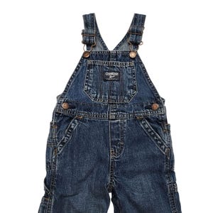 May include: Dark wash denim overalls with adjustable straps and metal hardware. The overalls feature a bib pocket with the "OshKosh B'gosh" logo patch. The garment has multiple pockets and bronze-coloured button closures. A classic children's garment.