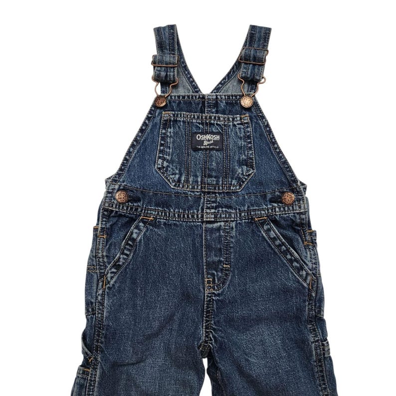 May include: Dark wash denim overalls with adjustable straps and metal hardware. The overalls feature a bib pocket with the "OshKosh B'gosh" logo patch. The garment has multiple pockets and bronze-coloured button closures. A classic children's garment.