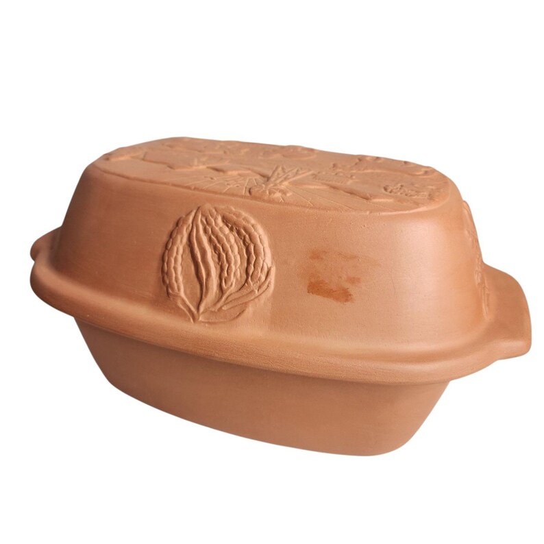 May include: A terracotta-colored oval-shaped baking dish with a lid. The lid and sides feature embossed floral and leaf designs. The dish has a smooth, unglazed finish and is designed for cooking.