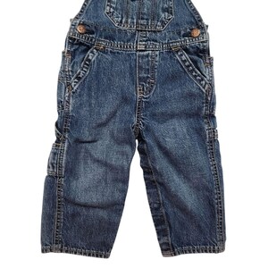 May include: Blue denim dungarees with adjustable straps, a bib pocket, and side pockets. The dungarees have metal button closures and are designed for children. The denim has a faded appearance.