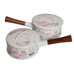 Op de afbeelding: Twee witte emaille steelpannen met deksels, versierd met roze, blauwe en gele bloemmotieven. Elke pan heeft een houten handvat en een wit deksel met een bijpassend bloemendesign. De handvatten zijn donkerbruin.