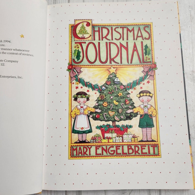 May include: A Christmas journal cover with a colorful illustration of a Christmas tree decorated with ornaments, candy canes, and a star. Two children stand on either side of the tree, holding hands. The text "CHRISTMAS JOURNAL" is written in a festive font above the illustration. The text "MARY ENGELBREIT" is written below the illustration.