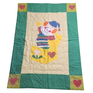 May include: A green and yellow patchwork quilt with a colorful appliqué of a clown in the center. The clown is wearing a blue hat and a red and white striped shirt. The quilt is edged with a green border.