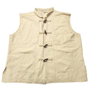 May include: A sleeveless cream-colored vest with a mandarin collar and five brown toggle closures. The vest has two front pockets and a single pocket on the right side.