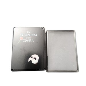 Könnte beinhalten: Eine schwarze Metallbox mit den Worten "The Phantom of the Opera" in weißer Schrift. Die Box zeigt eine weiße Maske und eine rote Rose. Die Box ist geöffnet und zeigt ein transparentes Kunststofffach.