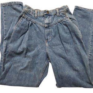May include: High-waisted blue denim jeans featuring a pleated front design, a button and zip fly closure, belt loops, and two side pockets. The jeans have a tapered leg silhouette and a medium wash finish with visible stitching details.