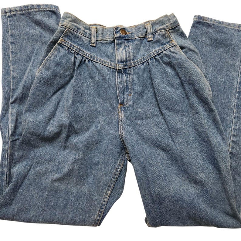 May include: High-waisted blue denim jeans featuring a pleated front design, a button and zip fly closure, belt loops, and two side pockets. The jeans have a tapered leg silhouette and a medium wash finish with visible stitching details.