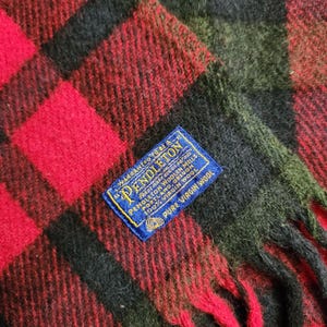 May include: A close-up shot of a red, black, and green plaid wool scarf. The scarf features a fringed edge and a blue label with the brand name "Pendleton" and "100% Virgin Wool". The plaid pattern is composed of large squares.