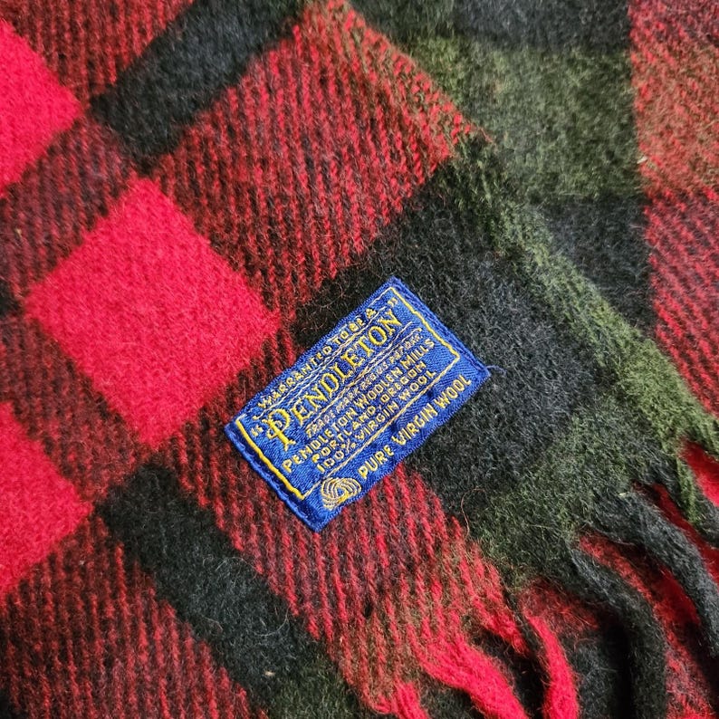 May include: A close-up shot of a red, black, and green plaid wool scarf. The scarf features a fringed edge and a blue label with the brand name "Pendleton" and "100% Virgin Wool". The plaid pattern is composed of large squares.