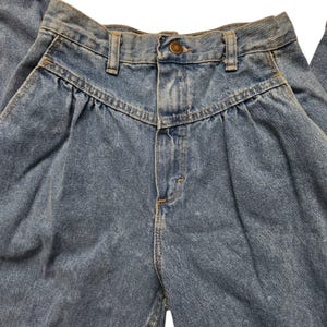 May include: High-waisted blue denim jeans featuring a pleated front design, a central metal button closure, a zip fly, and belt loops. The fabric has a medium wash finish with visible stitching details along the waistband and pockets.