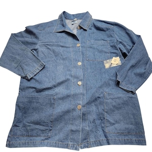 May include: A denim jacket with a collared neckline, long sleeves, and two front pockets. The jacket has a button-down closure and a small tag attached to the front pocket. The jacket is a medium blue color.