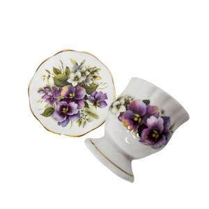 May include: A white porcelain egg cup and matching small saucer, both featuring a delicate floral pattern of purple pansies and green leaves. Each piece is finished with a thin gold trim along the scalloped edges, creating a cohesive set.