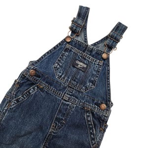May include: Dark blue denim overalls with metal clasps and adjustable straps. The overalls have a front pocket and the "OshKosh B'gosh" brand label. The garment has a classic, vintage style.