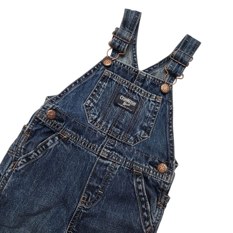 May include: Dark blue denim overalls with metal clasps and adjustable straps. The overalls have a front pocket and the "OshKosh B'gosh" brand label. The garment has a classic, vintage style.