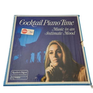 Könnte beinhalten: Ein Vintage-Vinyl-Schallplattenalbum mit dem Titel "Cocktail Piano Time" und dem Untertitel "Music in an Intimate Mood". Das Cover zeigt eine Frau mit langen Haaren und eine Klavierillustration auf blauem Hintergrund. Die Schallplatte ist in Kunststoff versiegelt.