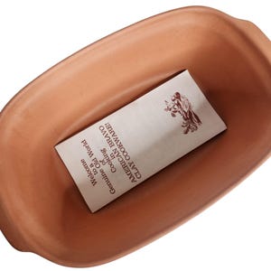 May include: A terracotta-colored rectangular baking dish with rounded edges. Inside the dish is a folded paper with text that reads "Welcome to a Genuine Old World Cooking in AMERICAN BRAVO CLAY COOKWARE!"