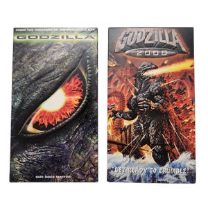 Può includere: Due videocassette VHS del film Godzilla. La prima presenta un primo piano dell'occhio di Godzilla con la scritta "SIZE DOES MATTER". La seconda videocassetta è intitolata "GODZILLA 2000" con Godzilla che sputa fuoco e la scritta "GET READY TO CRUMBLE!"