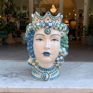 May include: A ceramic planter shaped like a woman's head with a crown, blue and green checkered pattern, and green grapes. The planter is white with blue and green details.
