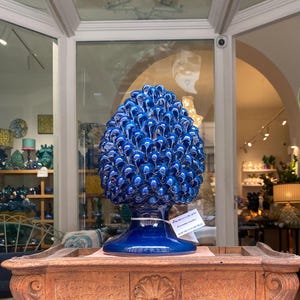 May include: A large, blue ceramic pinecone sculpture with a glossy finish. The sculpture is displayed on a wooden table in a shop setting. The pinecone is a vibrant shade of blue, with a detailed, textured surface.