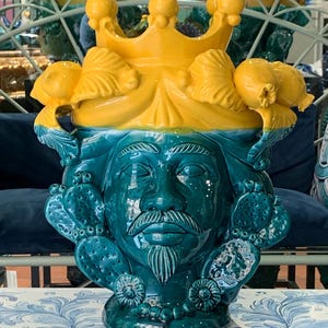 May include: A ceramic vase shaped like a man's head with a yellow crown and green details. The vase has a beard and mustache and is decorated with cactus-like plants.