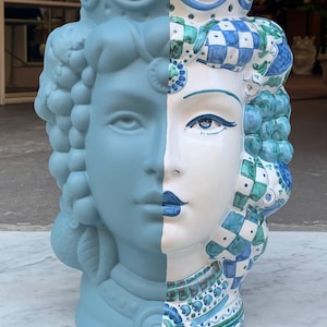 May include: A ceramic planter shaped like a woman's head, with one side painted in a light blue color and the other side decorated with a colorful pattern of blue, green, and white. The planter has a crown on top.