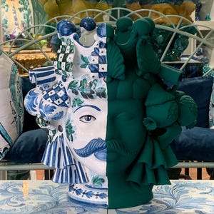 May include: A ceramic vase with a face design, half painted in blue and white and half in green. The vase has a crown and other decorative elements.