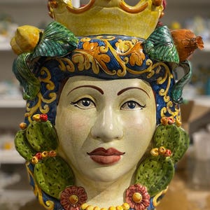 May include: A ceramic head vase with a yellow crown, green cactus, and red flowers. The vase is painted with blue, yellow, and green floral designs.
