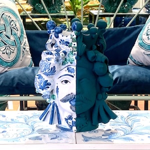 May include: A ceramic sculpture of a face, half painted in white with blue and green floral designs and the other half painted in a solid dark green. The sculpture is sitting on a table with a blue and white patterned surface.